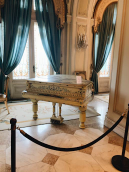 An elegant room with high ceilings and ornate architecture contains a decorative grand piano with intricate designs. The piano is positioned on an ornate rug, surrounded by polished marble flooring. Rich teal curtains hang beside decorative glass doors, and there is a crystal chandelier mounted on the wall.