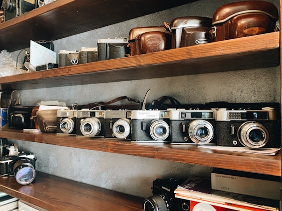 A shelf filled with a collection of vintage cameras and their leather cases. The wooden shelves are neatly organized with cameras of varying designs, predominantly classic models with metallic and black finishes. The ambiance is nostalgic, evoking a sense of appreciation for photography's history.