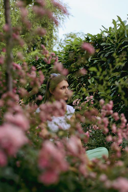 A serene woman sitting peacefully in a sunlit garden surrounded by soft greenery and blush pink flowers.