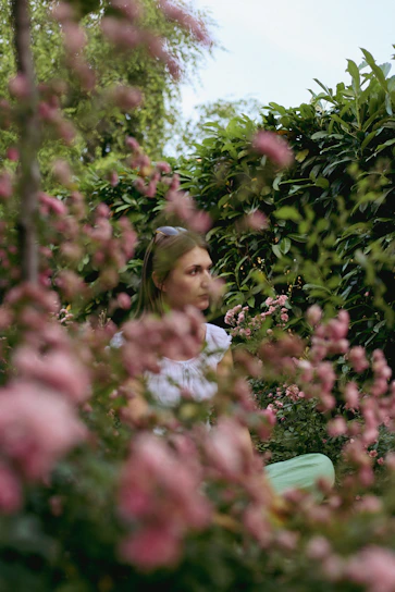 A serene woman sitting peacefully in a sunlit garden surrounded by soft greenery and blush pink flowers.
