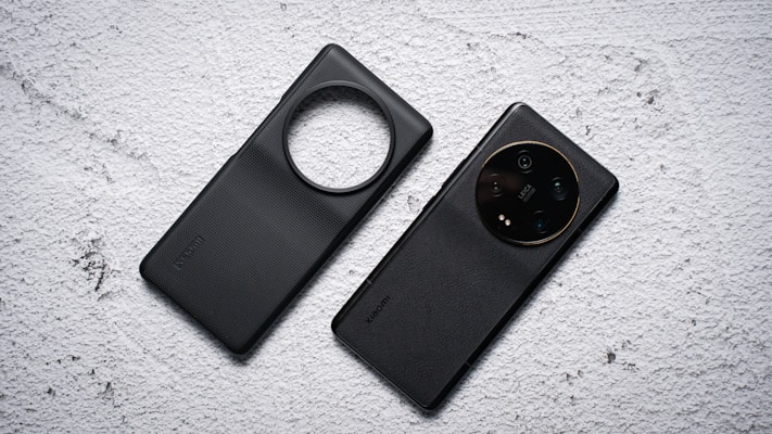 Two black mobile phone cases are placed side by side on a textured light gray surface. One case has a circular cutout for the camera, while the other is designed to highlight the camera module with lenses visible.