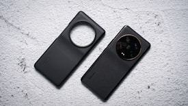 Two black mobile phone cases are placed side by side on a textured light gray surface. One case has a circular cutout for the camera, while the other is designed to highlight the camera module with lenses visible.