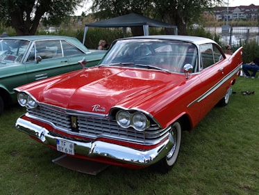 A classic red Plymouth car with polished chrome details and large round headlights is parked on a grassy lawn. Nearby, an older light green car is visible, and people are seated under a canopy in the background.
