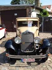 A vintage car with a beige and black color scheme is parked on cobblestone near a brick garage. The car displays a classic grille and round headlights. Surrounding the car are residential buildings and greenery.