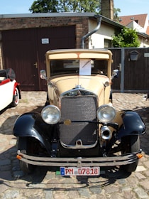 A vintage car with a beige and black color scheme is parked on cobblestone near a brick garage. The car displays a classic grille and round headlights. Surrounding the car are residential buildings and greenery.