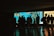 A group of silhouetted people stand in front of an illuminated aquarium tank, observing various sea creatures swimming within a large, blue-lit enclosure. The ambiance is dark, with the primary light source coming from the tank, casting shadows that accentuate the figures of the observers.
