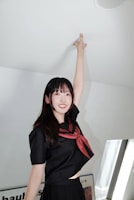 A young woman in a dark school uniform with a red scarf is reaching up toward the ceiling, standing in a well-lit room. The room has soft white and gray tones with some framed artwork visible in the background.