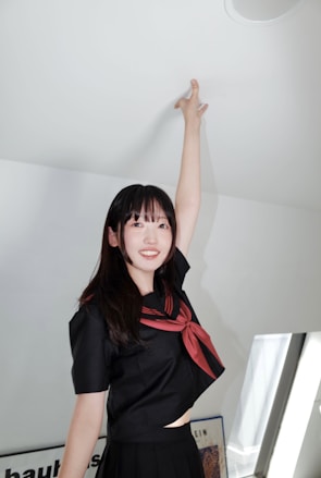 A young woman in a dark school uniform with a red scarf is reaching up toward the ceiling, standing in a well-lit room. The room has soft white and gray tones with some framed artwork visible in the background.