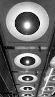 Circular ceiling lights are arranged in a linear fashion, emitting bright light. The scene includes a reflective glass surface on the right, and the lights cast geometric patterns and shadows. The image is monochrome, enhancing the contrasts and shapes.