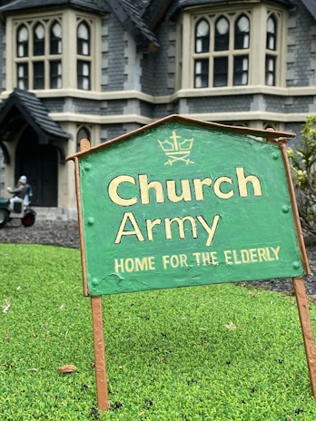 A green sign with the text 'Church Army Home for the Elderly' is prominently displayed on a patch of grass. In the background, a large stone building with gothic-style windows can be seen, suggesting an old-style architectural design. A figure in the background appears to be tending to the garden or performing some task near the building.