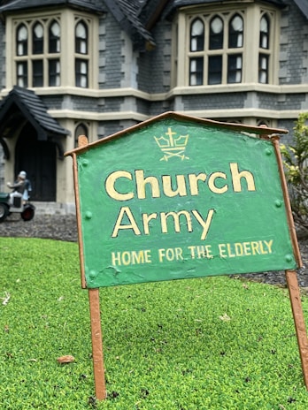 A green sign with the text 'Church Army Home for the Elderly' is prominently displayed on a patch of grass. In the background, a large stone building with gothic-style windows can be seen, suggesting an old-style architectural design. A figure in the background appears to be tending to the garden or performing some task near the building.