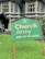 A green sign with the text 'Church Army Home for the Elderly' is prominently displayed on a patch of grass. In the background, a large stone building with gothic-style windows can be seen, suggesting an old-style architectural design. A figure in the background appears to be tending to the garden or performing some task near the building.