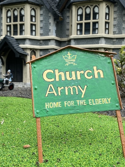 A green sign with the text 'Church Army Home for the Elderly' is prominently displayed on a patch of grass. In the background, a large stone building with gothic-style windows can be seen, suggesting an old-style architectural design. A figure in the background appears to be tending to the garden or performing some task near the building.