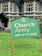 A green sign with the text 'Church Army Home for the Elderly' is prominently displayed on a patch of grass. In the background, a large stone building with gothic-style windows can be seen, suggesting an old-style architectural design. A figure in the background appears to be tending to the garden or performing some task near the building.