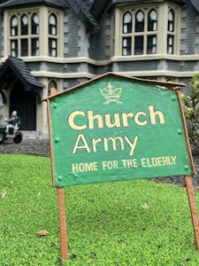 A green sign with the text 'Church Army Home for the Elderly' is prominently displayed on a patch of grass. In the background, a large stone building with gothic-style windows can be seen, suggesting an old-style architectural design. A figure in the background appears to be tending to the garden or performing some task near the building.