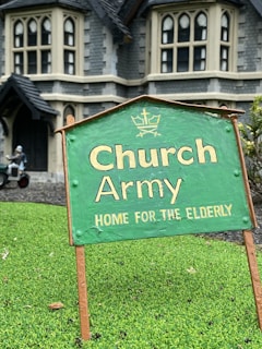 A green sign with the text 'Church Army Home for the Elderly' is prominently displayed on a patch of grass. In the background, a large stone building with gothic-style windows can be seen, suggesting an old-style architectural design. A figure in the background appears to be tending to the garden or performing some task near the building.