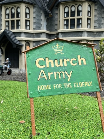 A green sign with the text 'Church Army Home for the Elderly' is prominently displayed on a patch of grass. In the background, a large stone building with gothic-style windows can be seen, suggesting an old-style architectural design. A figure in the background appears to be tending to the garden or performing some task near the building.