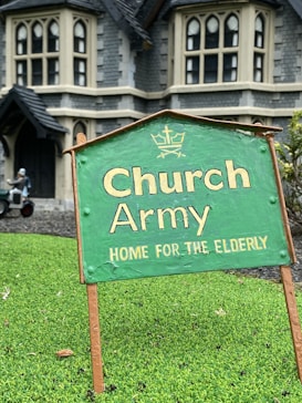 A green sign with the text 'Church Army Home for the Elderly' is prominently displayed on a patch of grass. In the background, a large stone building with gothic-style windows can be seen, suggesting an old-style architectural design. A figure in the background appears to be tending to the garden or performing some task near the building.