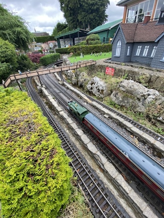 A model railway setup featuring a miniature green train moving on tracks surrounded by lush greenery and rocky terrain. There is a small bridge over the railway and several buildings, including a grey house with blue windows, in the background. The setting appears to be an outdoor miniature park with well-maintained landscaping.