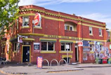 A two-story brick building with yellow trim features prominently. The building, identified as the Union Club Hotel, has a vintage look with a red and green awning above the entrance. A red circular sign for Carlton Draught is displayed prominently. Outside, there are metal bike racks and garbage bins, along with some seating under the shade of a nearby tree.