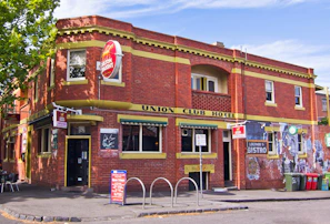 A two-story brick building with yellow trim features prominently. The building, identified as the Union Club Hotel, has a vintage look with a red and green awning above the entrance. A red circular sign for Carlton Draught is displayed prominently. Outside, there are metal bike racks and garbage bins, along with some seating under the shade of a nearby tree.