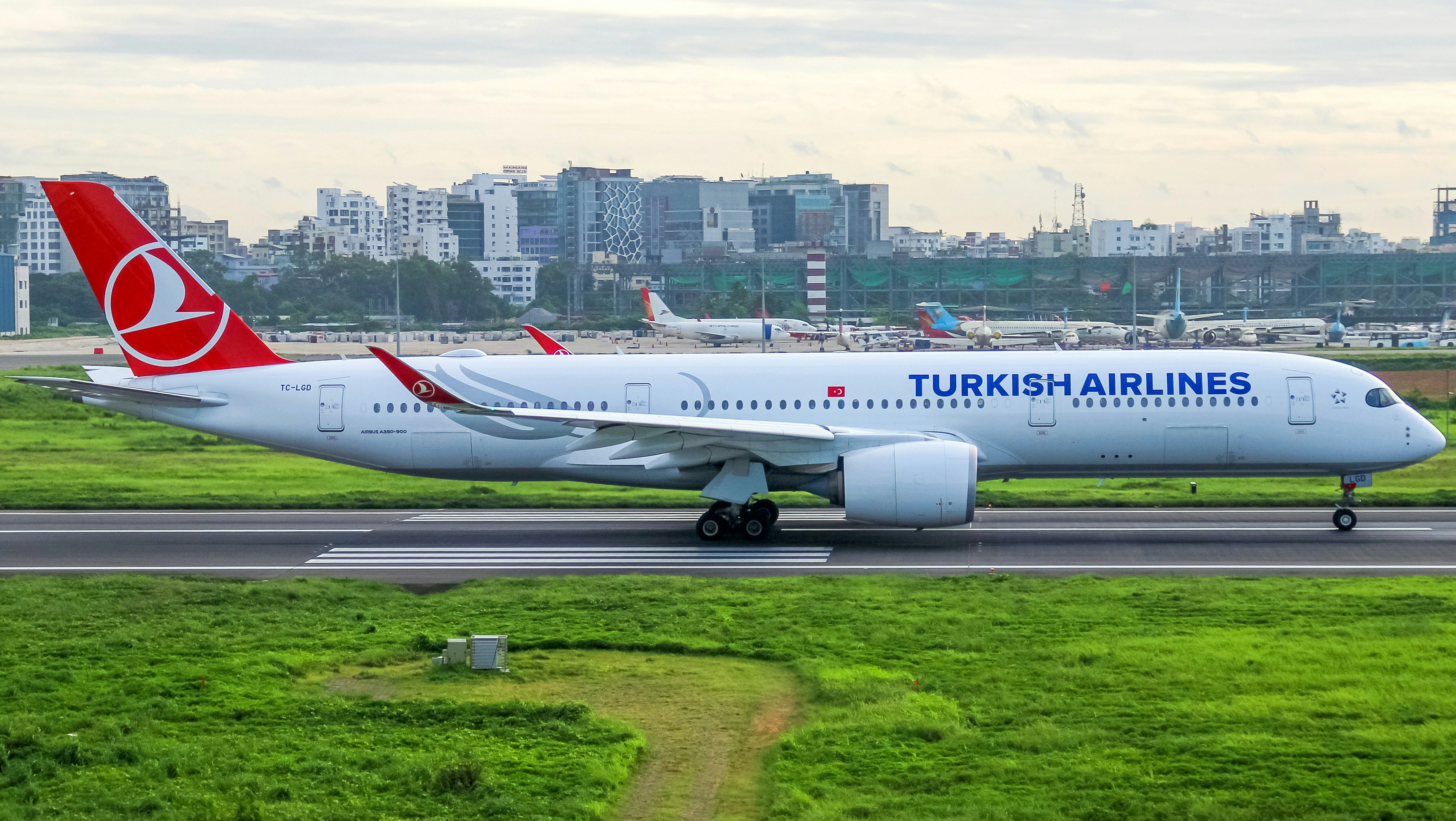Photo of a modern airplane flying low over green fields with mountains in background and partly cloudy sky