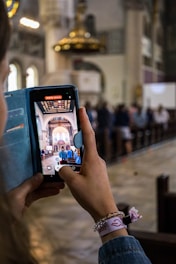 A person is holding a smartphone, capturing a video inside a large, ornate church. The screen displays the interior of the church with arched ceilings, intricate designs, and people seated in pews.