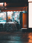 A small convenience store with bright internal lighting contrasts with the dimly lit exterior. Various colorful products are displayed, including snacks, drinks, and magazines. A person is inside, partially obscured by the shelves and merchandise.