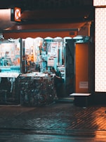 A small convenience store with bright internal lighting contrasts with the dimly lit exterior. Various colorful products are displayed, including snacks, drinks, and magazines. A person is inside, partially obscured by the shelves and merchandise.