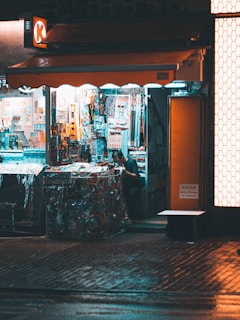 A small convenience store with bright internal lighting contrasts with the dimly lit exterior. Various colorful products are displayed, including snacks, drinks, and magazines. A person is inside, partially obscured by the shelves and merchandise.