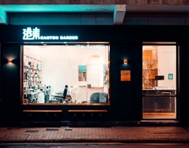A barbershop with a dimly lit exterior and illuminated interior. Through the large window, barber chairs and shelves stocked with products are visible. The shop has a modern and sleek design, with dark walls and warm lighting inside. Signage is displayed above the entrance, and there is a small bench outside on the pavement.