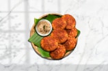 A plate with crispy, golden-brown patties on a banana leaf, accompanied by a small bowl of creamy coconut chutney. The patties are arranged neatly in a wicker basket set against a marble surface with a shadow pattern adding depth.