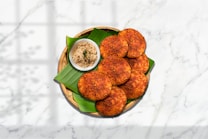 A plate with crispy, golden-brown patties on a banana leaf, accompanied by a small bowl of creamy coconut chutney. The patties are arranged neatly in a wicker basket set against a marble surface with a shadow pattern adding depth.
