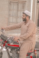 A man wearing traditional South Asian attire and a cap is seated on a red and black motorcycle. The background features a rustic brick wall and a wooden door, giving a rural or small-town ambiance.