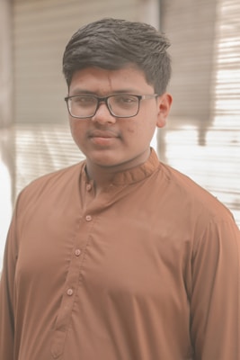 A young person wearing glasses and a traditional brown kurta, standing indoors with soft lighting. The background features window blinds that diffuse light across the scene.