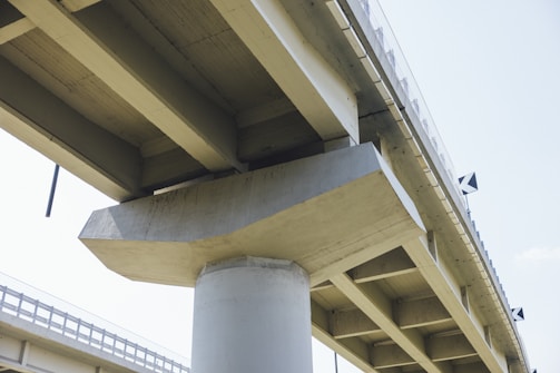 A close-up view of a concrete bridge with large supporting beams and columns. The structure appears robust and is highlighted against a pale sky. The geometric lines of the bridge create a sense of strength and stability.
