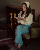 A woman with long dark hair, wearing a white shirt and blue jeans, sits on an armchair while reading a book. The setting features a cozy room with two lit candles on a table beside her, adding a warm ambiance. A patterned rug is partially visible on the wooden floor.