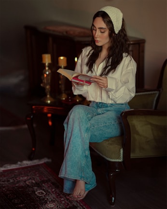 A woman with long dark hair, wearing a white shirt and blue jeans, sits on an armchair while reading a book. The setting features a cozy room with two lit candles on a table beside her, adding a warm ambiance. A patterned rug is partially visible on the wooden floor.