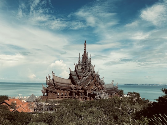 An intricately designed wooden temple sits majestically by the sea, its elaborate spires and carvings reaching towards the sky. The structure is surrounded by lush greenery, and the ocean stretches out behind it, meeting a partly cloudy sky.