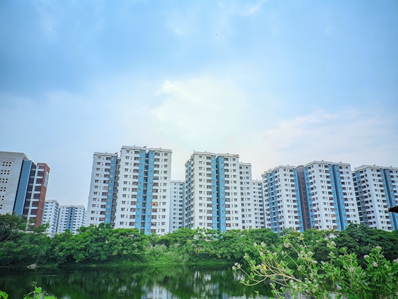A row of modern high-rise apartment buildings is set against a slightly overcast sky. In front of the buildings, there is a lush green landscape with various trees and a calm body of water reflecting part of the skyline.