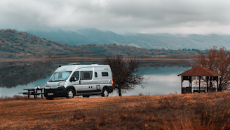 A camper van is parked near a calm lake, with a picnic table and a gazebo nearby amidst a tranquil landscape. Rolling hills and mountains form the backdrop, partially covered by a layer of clouds. The surroundings are bathed in autumn hues, contributing to a serene atmosphere.