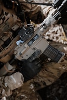 A person wearing camouflage clothing is holding a tan-colored rifle with a black handguard and a transparent magazine. The gun has '300 BLK' markings on it, and the person is wearing black tactical gloves. There is a background of natural foliage.