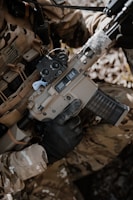 A person wearing camouflage clothing is holding a tan-colored rifle with a black handguard and a transparent magazine. The gun has '300 BLK' markings on it, and the person is wearing black tactical gloves. There is a background of natural foliage.