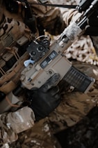 A person wearing camouflage clothing is holding a tan-colored rifle with a black handguard and a transparent magazine. The gun has '300 BLK' markings on it, and the person is wearing black tactical gloves. There is a background of natural foliage.