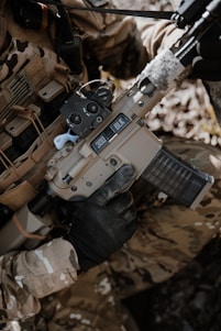 A person wearing camouflage clothing is holding a tan-colored rifle with a black handguard and a transparent magazine. The gun has '300 BLK' markings on it, and the person is wearing black tactical gloves. There is a background of natural foliage.