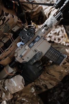 A person wearing camouflage clothing is holding a tan-colored rifle with a black handguard and a transparent magazine. The gun has '300 BLK' markings on it, and the person is wearing black tactical gloves. There is a background of natural foliage.
