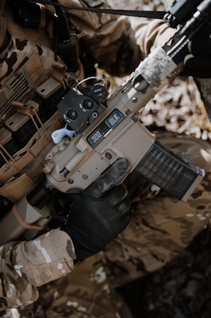 A person wearing camouflage clothing is holding a tan-colored rifle with a black handguard and a transparent magazine. The gun has '300 BLK' markings on it, and the person is wearing black tactical gloves. There is a background of natural foliage.