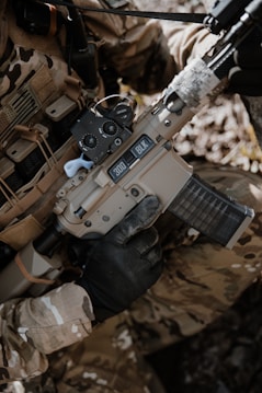 A person wearing camouflage clothing is holding a tan-colored rifle with a black handguard and a transparent magazine. The gun has '300 BLK' markings on it, and the person is wearing black tactical gloves. There is a background of natural foliage.