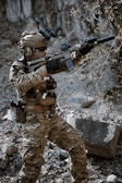 A person wearing full tactical military gear is holding a rifle equipped with a scope and suppressor. The individual is dressed in camouflage clothing and appears to be standing in a rocky, outdoor setting with a focused stance.