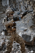 A person wearing full tactical military gear is holding a rifle equipped with a scope and suppressor. The individual is dressed in camouflage clothing and appears to be standing in a rocky, outdoor setting with a focused stance.